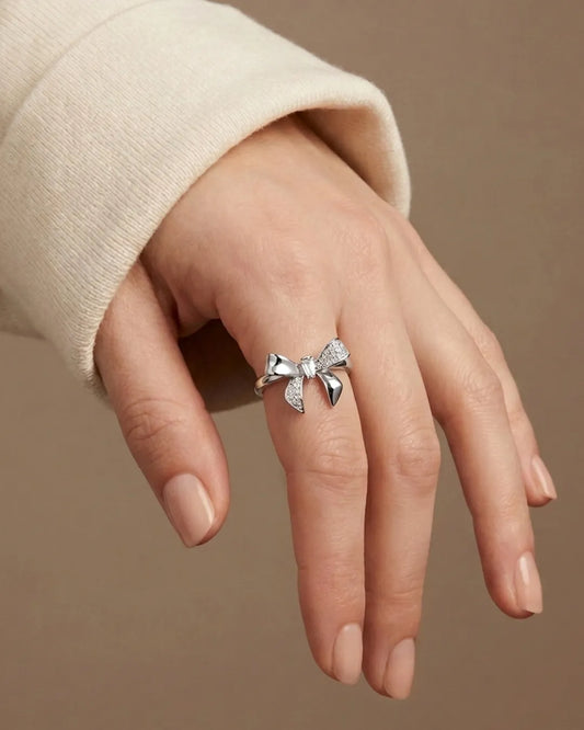 Silver bow-shaped ring on a hand against a beige background