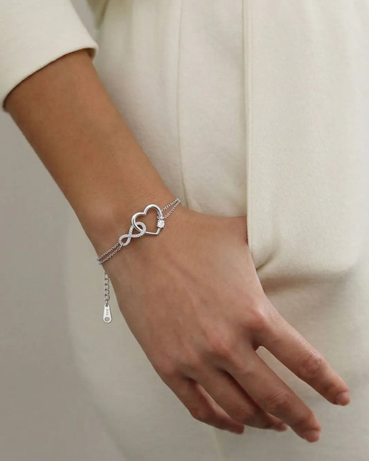 Silver bracelet with heart and infinity symbol on a wrist against a neutral background