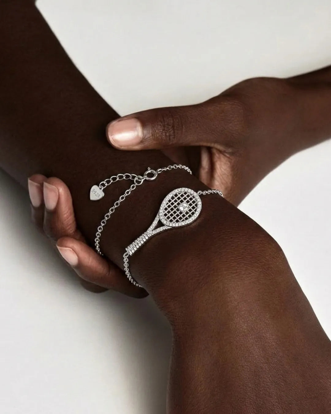 Two hands wearing silver bracelet with tennis racket design on a white background